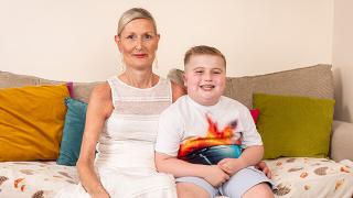 A smiling woman in a white dress sits on a sofa beside a happy young boy in a colorful T-shirt, both looking at the camera