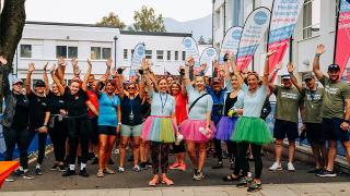 Group of cheerful people in colorful outfits and tutus raise their hands at a fundraising event outside a building with banners behind them.