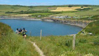Two hikers ascend a grassy coastal path with trekking poles, overlooking a tranquil bay surrounded by cliffs. The water below is dotted with kayakers, and a car park with vehicles is visible on the clifftop opposite, under a bright, sunny sky