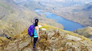 A woman with a purple backpack stands on a rocky mountain ridge, looking out over a vast landscape of rugged peaks and a deep blue lake below. The scene is expansive and dramatic, with a clear view of winding trails and the distant terrain.