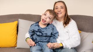A woman and a young boy with a disability sit closely together on a gray couch with yellow and gray pillows. The woman wears a white sweater and jeans, smiling warmly, while the boy, in a blue tie-dye shirt and jeans, leans against her, also smiling.