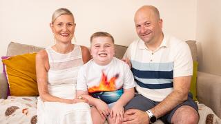 Smiling boy sits on a sofa between a woman in a white dress and a man in a striped polo, posing happily together indoors.