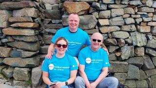 Three people in matching blue "mountain rescue for charity" shirts pose by a stone structure on a rocky mountain summit.