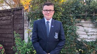 Luke wearing glasses and a navy school uniform stands in a garden, smiling, with greenery and wooden fences in the background.