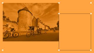 Cyclists ride past a historic stone tower and buildings on a sunny day, captured through an orange-tinted filter.