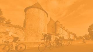 Cyclists ride past a historic stone tower and buildings on a sunny day, captured through an orange-tinted filter.