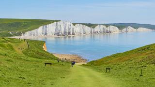 Walkers approach the beach near the iconic white chalk cliffs of the Seven Sisters, bordered by rolling green hills and calm blue sea.