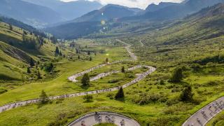 High-angle shot of hundreds of cyclists navigating a winding mountain road with sharp switchbacks through lush green valleys under a partly cloudy sky.