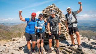 team of four stand on the summit of the old man of coniston with clear skies and views of the surrounding mountain range