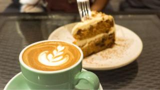 Close-up of a latte with leaf art and a slice of cake on a white plate, blurred in the background.