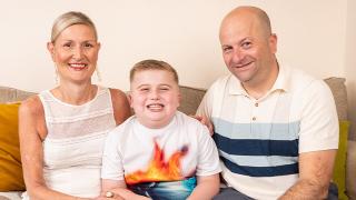 Digby with Mum and Dad -  Gemini said A smiling man and woman sit on a sofa with a young boy, who is wearing a colorful t-shirt.