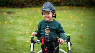 A picutre of Barnaby, a young boy in a green bear sweater and cap smiles while using a supportive walking frame in a grassy garden with fallen leaves.