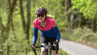 A close-up panoramic view of a female cyclist wearing a helmet, sunglasses, and a pink and blue Fdx jersey with trees behind her.