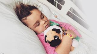 A child, Lilly-May, sleeping in a hospital bed with a visible head incision, holding a stuffed pug puppy toy closely.