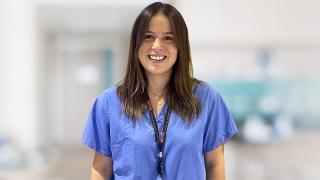 Dr. Eva Ioannidou, a smiling woman with brown hair wears blue medical scrubs and a UCL lanyard in a blurred hospital hallway.