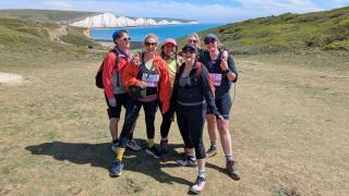 A group of six smiling female hikers posing together on a grassy coastal trail, with a vibrant blue sea and the iconic white chalk cliffs of the Seven Sisters in the background on a sunny day.