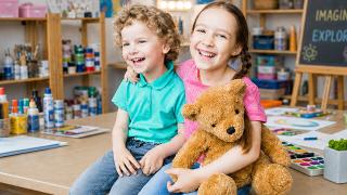 Two smiling children embrace while sitting on a table in a classroom, with the girl holding a plush teddy bear.