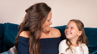 A mother and her young daughter sit on a teal couch, smiling and looking happily at each other.