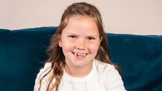 Hattie, a smiling young girl with light brown hair wearing a white top sits in front of a blue velvet couch.
