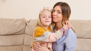 A young blonde girl wearing a floral headband warmly embraces her mother while sitting together on a beige couch.