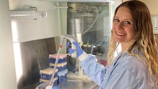 A smiling female researcher in a lab coat and gloves using a pipette inside a laboratory fume hood.