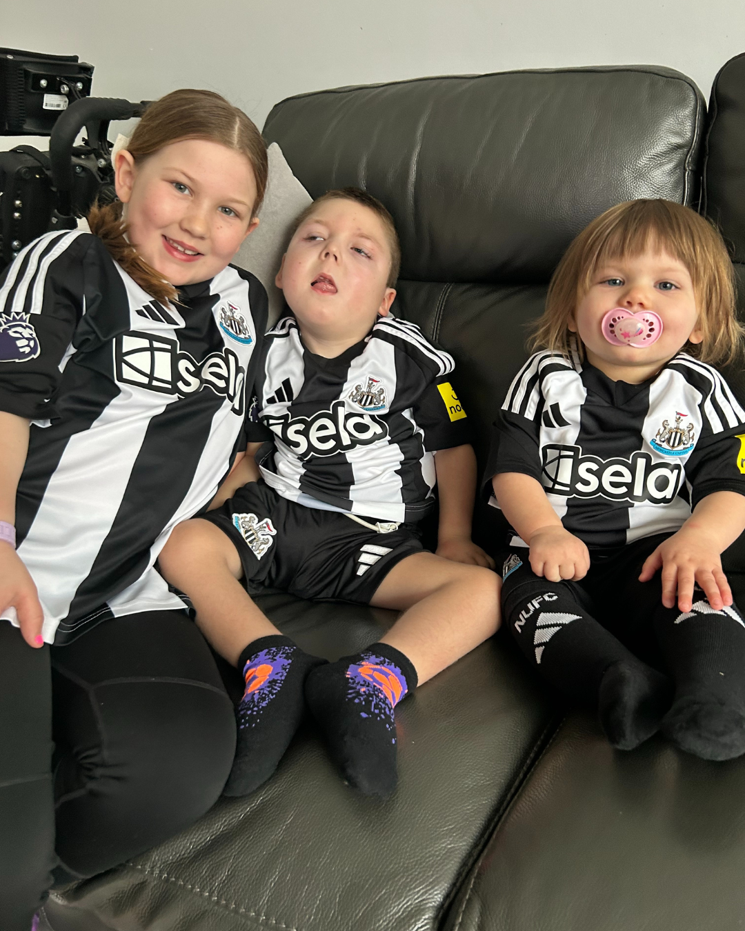 Three young children sit on a black leather couch wearing matching Newcastle United football jerseys, smiling and relaxed.